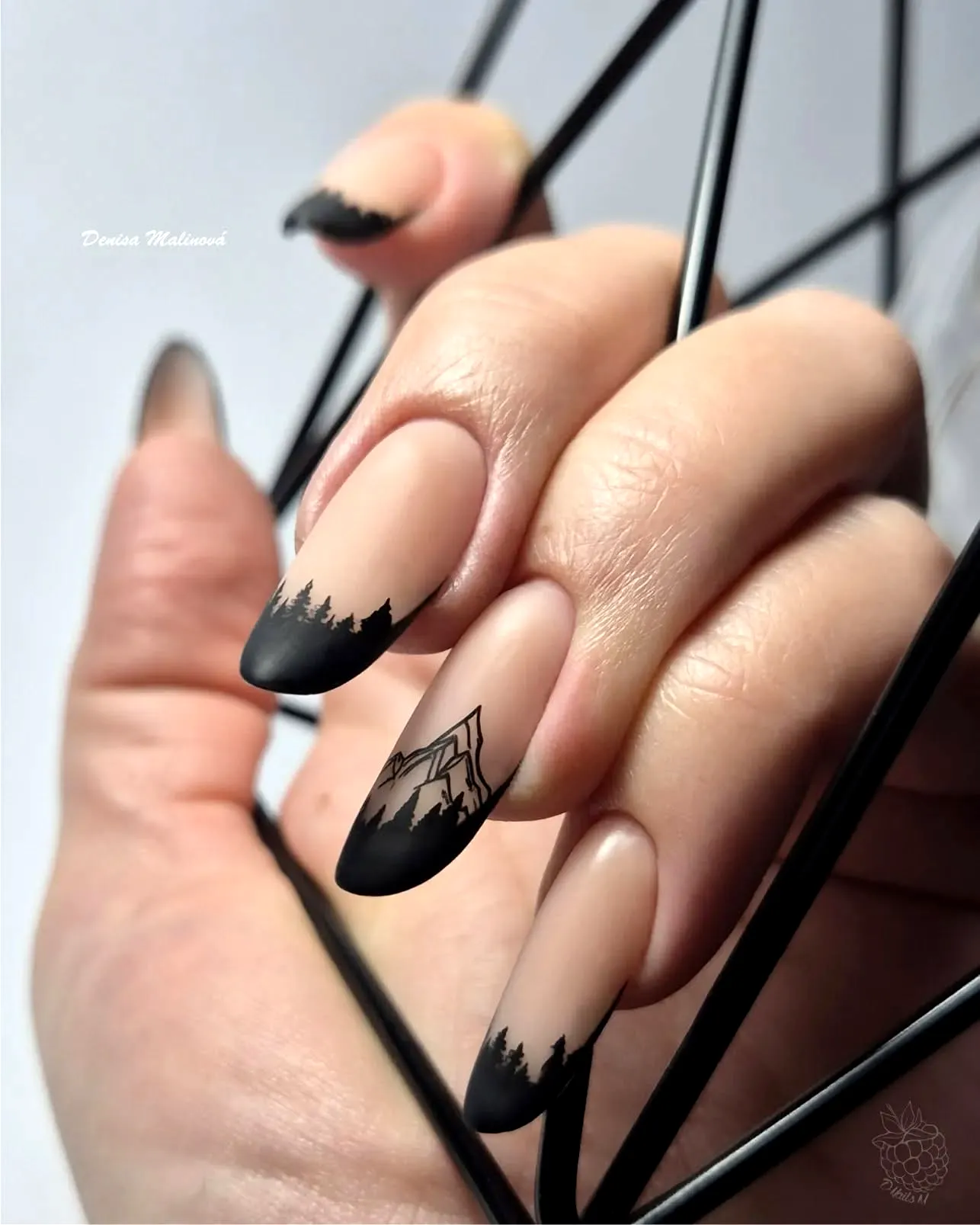 Chic stiletto mountain nails with nude base and glossy black French tips featuring nature-inspired silhouettes