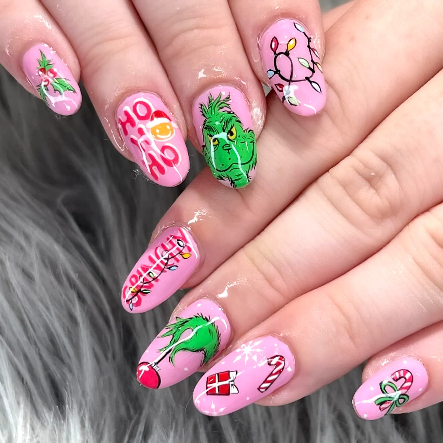Cute oval Grinch nails with pink background featuring cartoonish Grinch artwork Christmas lights candy canes and gifts in a whimsical nail art style