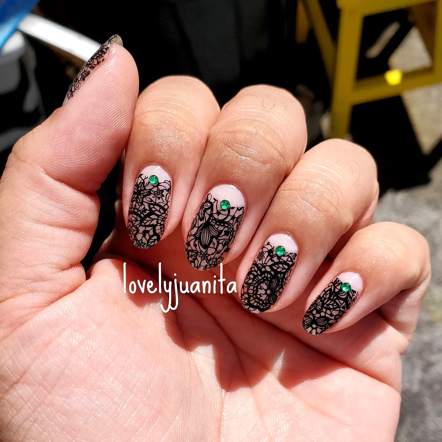 Elegant Almond Lace Nails with Black Floral Design on Nude Base and Green Gemstone Accents