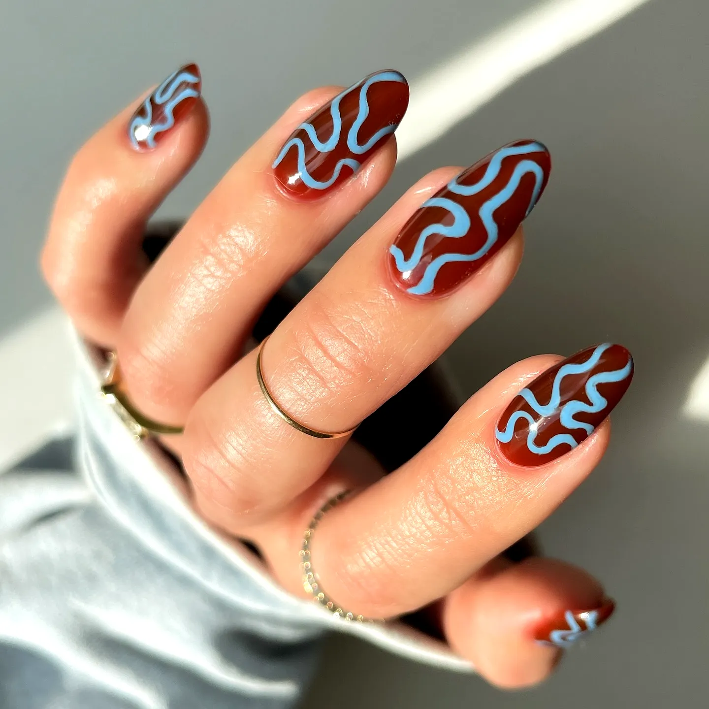 Elegant almond shaped swirl nail designs with glossy deep reddish-brown and light blue wavy lines