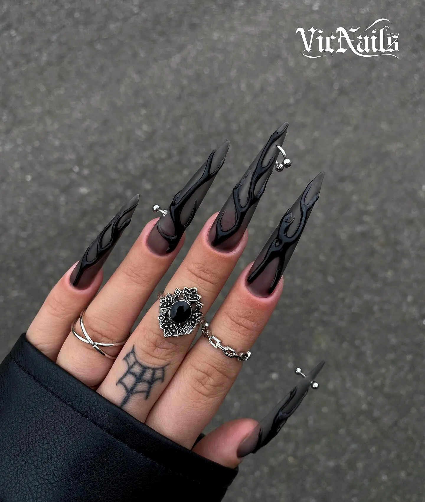 Gothic matte black stiletto nails with intricate swirling patterns and silver accents